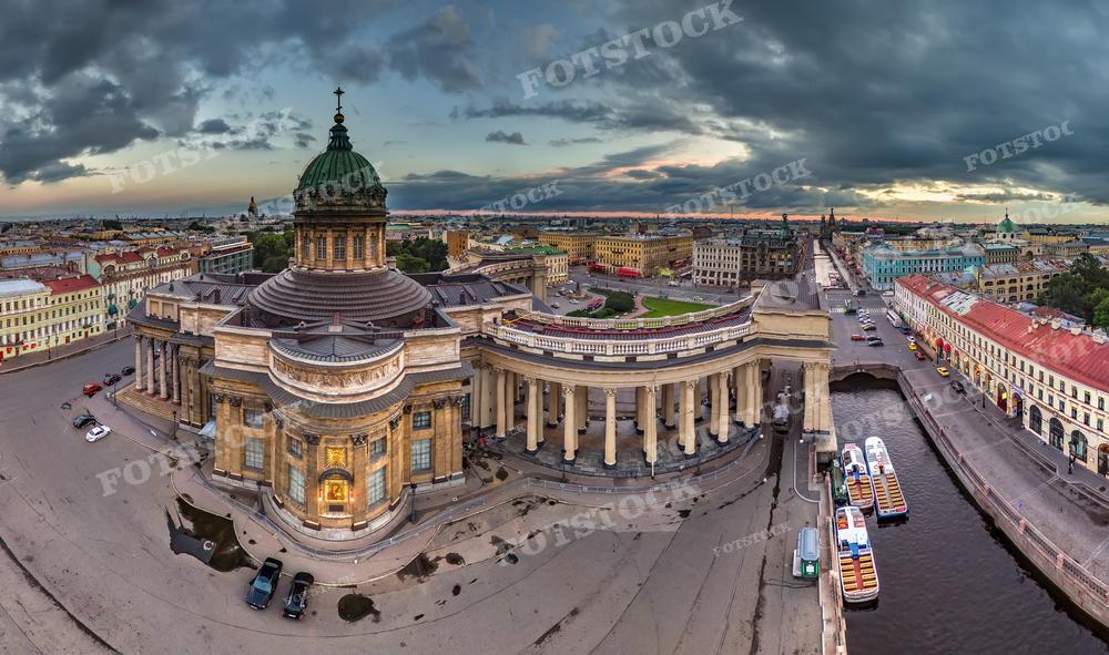 Эта панорамная фотография захватывает Казанский собор в Санкт-Петербурге. Изображение выполнено в сумерки или на рассвете, что видно по освещению фасада собора и тёмных облаках на небе, на фоне которых проступают светлые просветы. Рядом с собором видна набережная с причалами и туристическими корабликами. Такая фотография может быть использована в религиозной литературе, исторических изданиях, туристических брошюрах или как элемент дизайна интерьера.